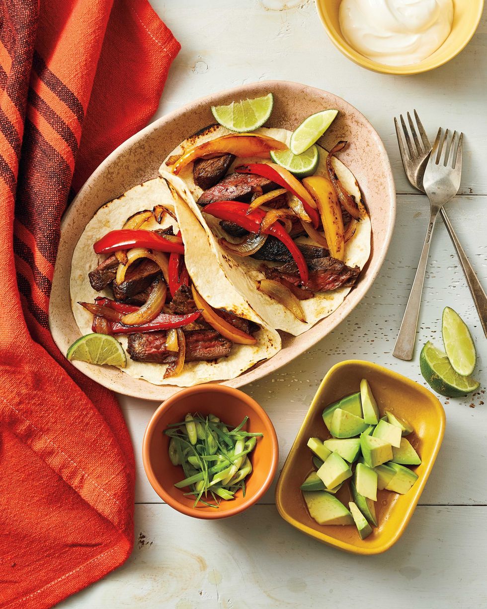 air fryer fajitas with lime wedges in a light tan colored oval bowl with small bowls of sliced green onion diced avocados and sour cream